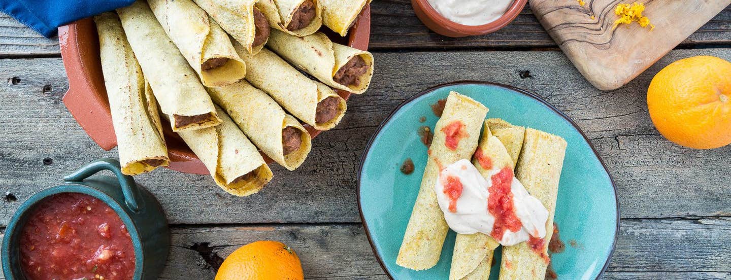 A serving bowl full of Orange Black Bean Taquitos, with three taquitos on a blue serving plate, topped with salsa and tofu cream