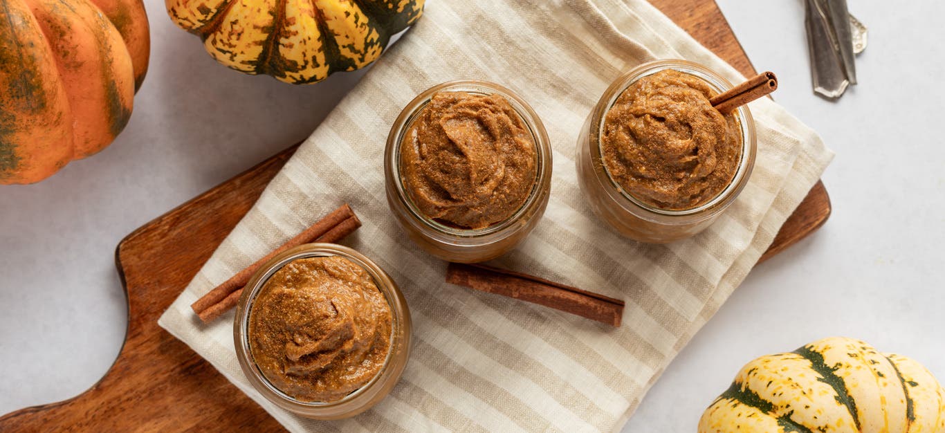 A top-down shot of three jars of almost-instant pumpkin pudding, with two sticks of cinnamon and some pumpkins lying beside the glasses, an easy vegan dessert recipe by Chef AJ