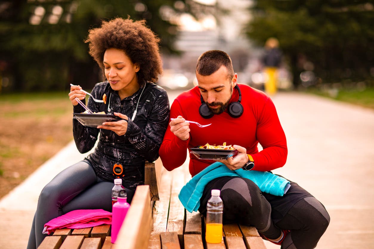 Two vegan athletes, a woman and man who appear to be in their early 30s, sit side by side on benches eating a healthy lunch