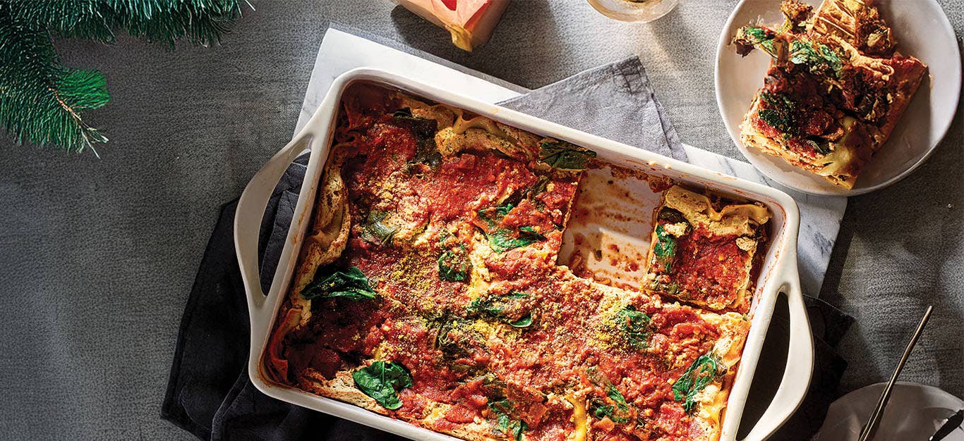 A white casserole dish of Spinach Lasagna with one slice removed and served on a white plate to the side. Set in a dimly lit grey setting.