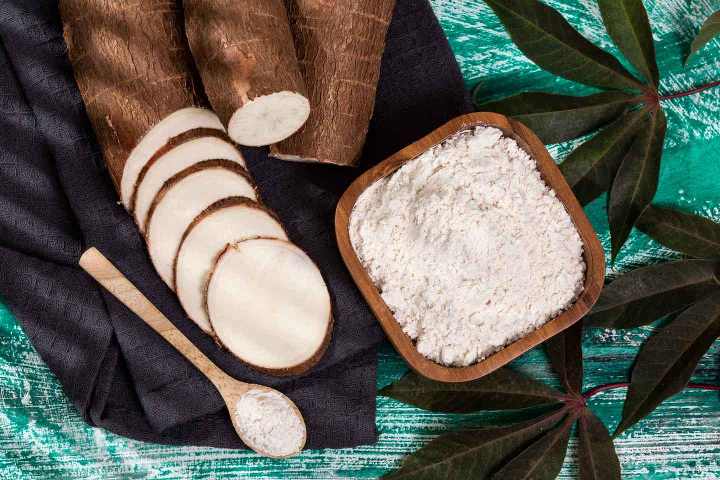 raw cassava (yuca) root shown sliced next to a bowl of cassava starch on a blue wooden table with leaves to the side