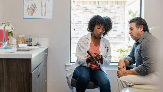 A doctor talks to a patient, who sits upright on the examination table, looking at a tablet that the doctor is holding up