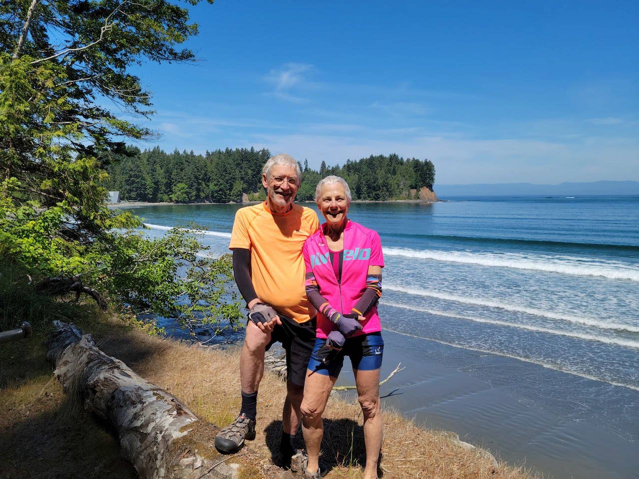 Janet Anspach Rickey and her husband stand on the shore of a lake, wearing cycling outfits, looking healthy after adopting a WFPB sos-free diet for weight loss and heart health