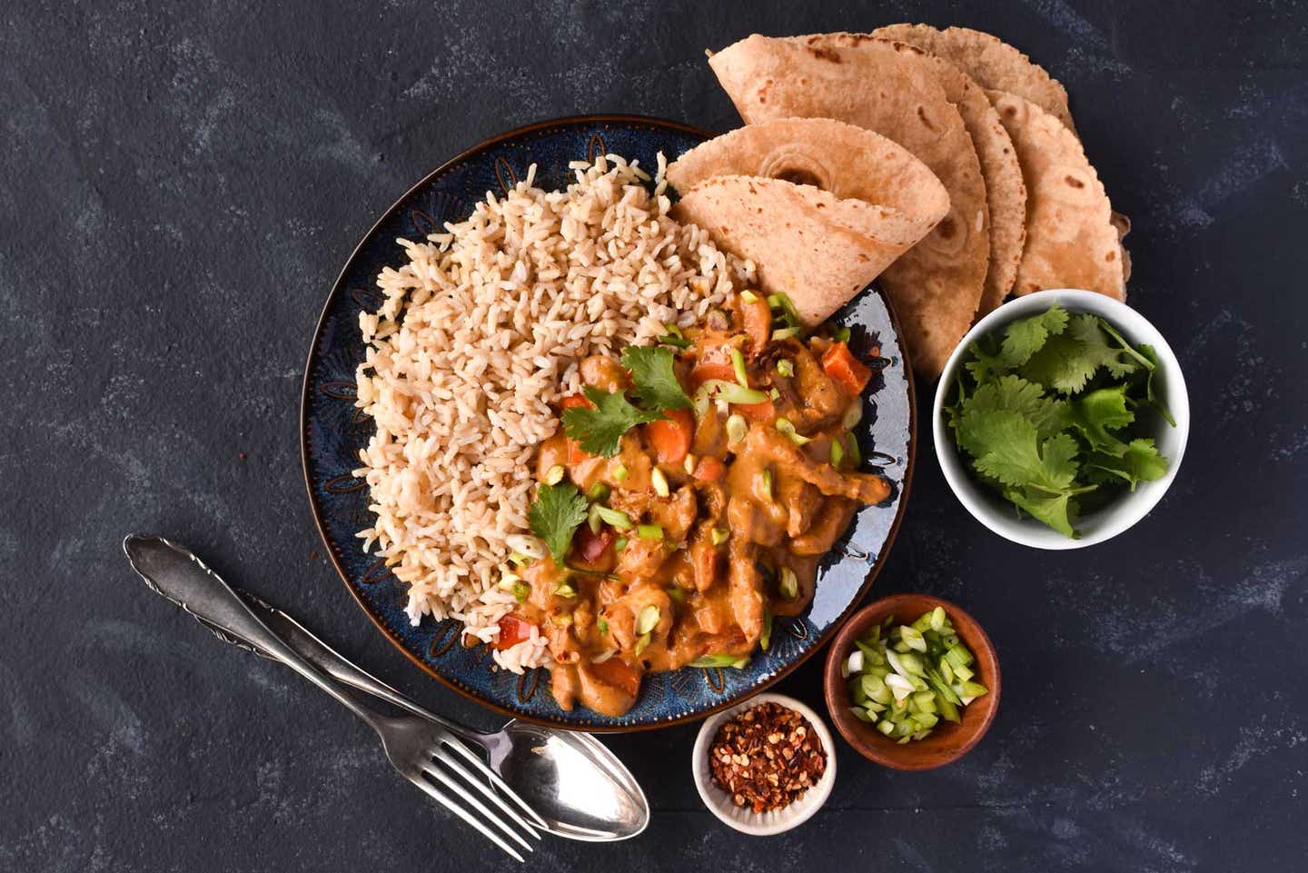 Vegetable Tikka Masala in a large black bowl surrounded by small bowls of cilantro, scallions, and chili flakes