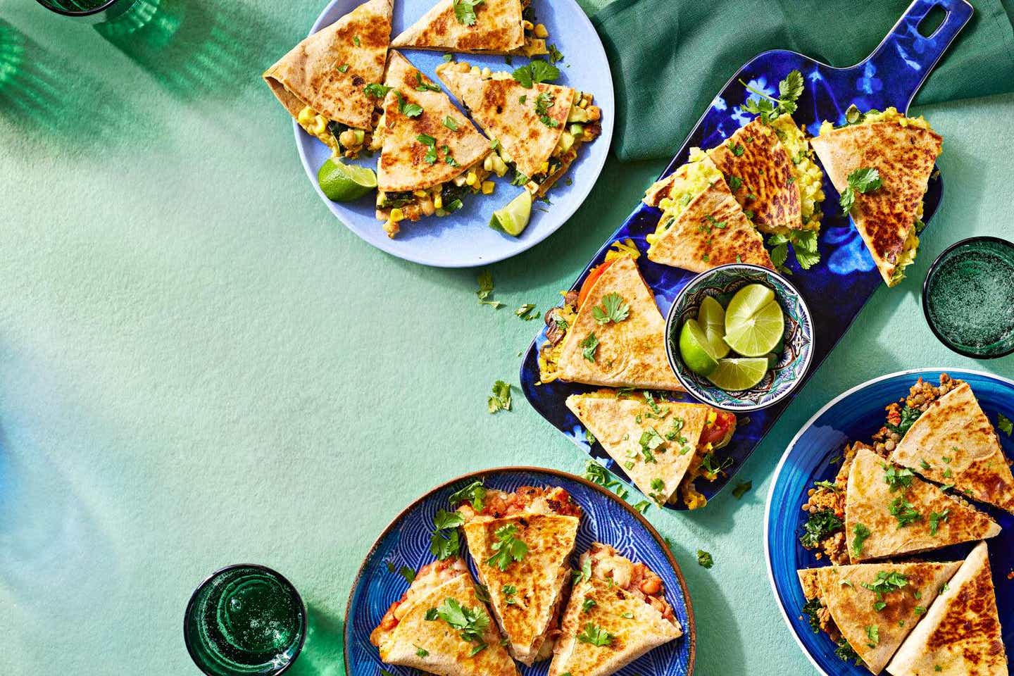 An overhead shot of five different types of vegan quesadillas, on plates on a light blue table