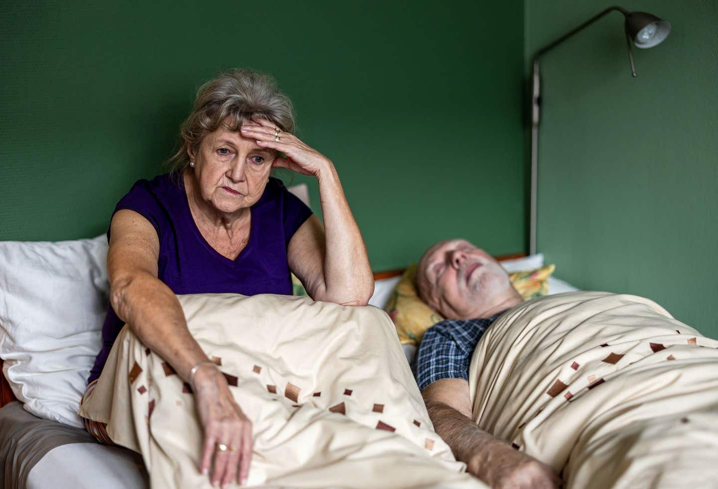 An older white woman sits up in bed, unable to sleep, while her husband lies snoring next to her