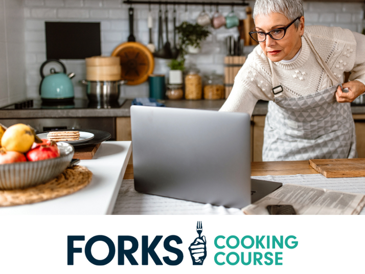 Older woman standing in her kitchen wearing an apron, leaning over, looking at her laptop