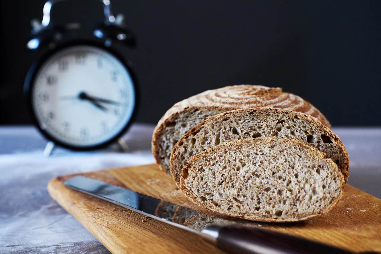 A loaf of sliced whole wheat bread sits on a wooden cutting board with an old alarm clock in the background