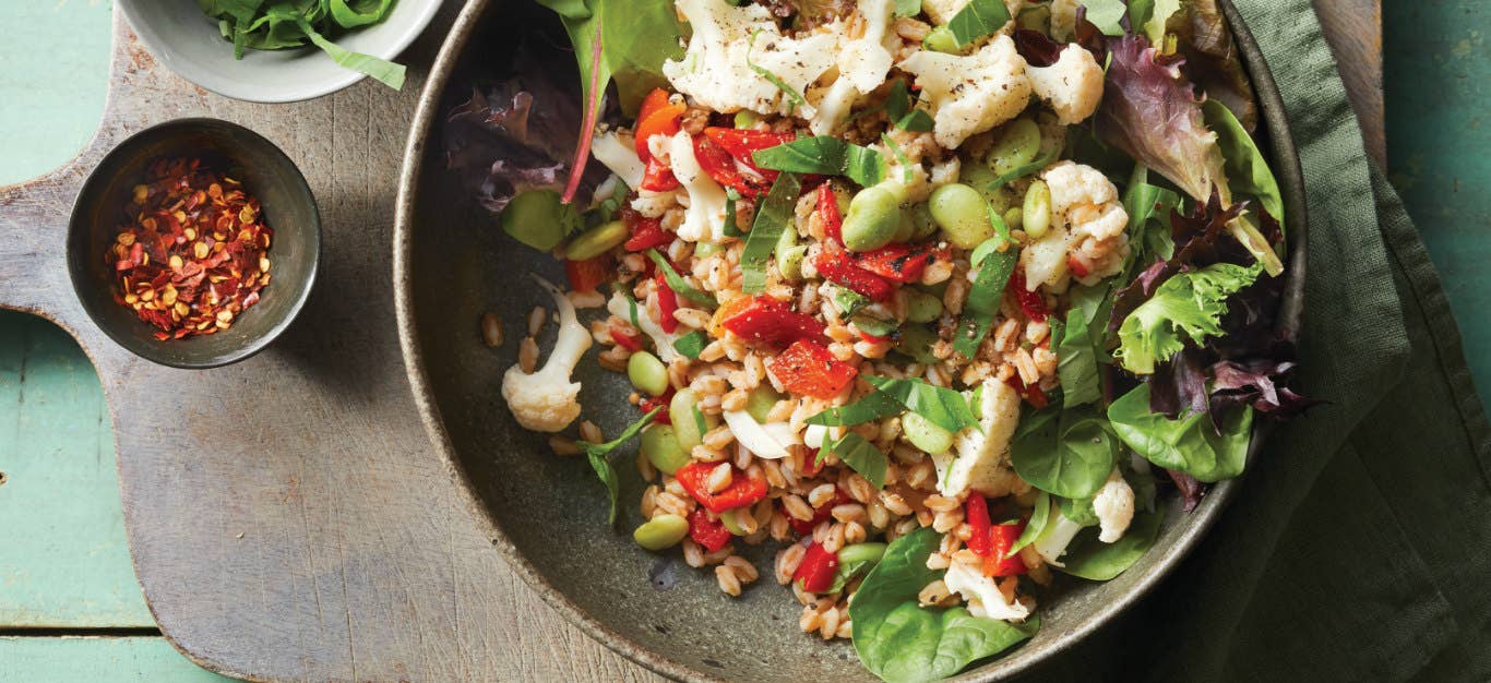 Pickled Cauliflower and Farro Salad in a brown earthy serving bowl on a wooden chopping board next to a small bowl of crushed red pepper