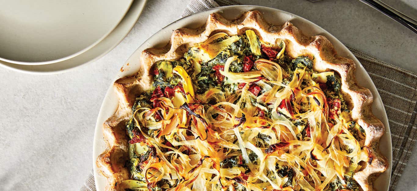 Quiche Florentine with Artichoke Hearts and Sun-Dried Tomatoes on a white plate on a white tablecloth with a beige and white striped napkin and a couple of empty plates