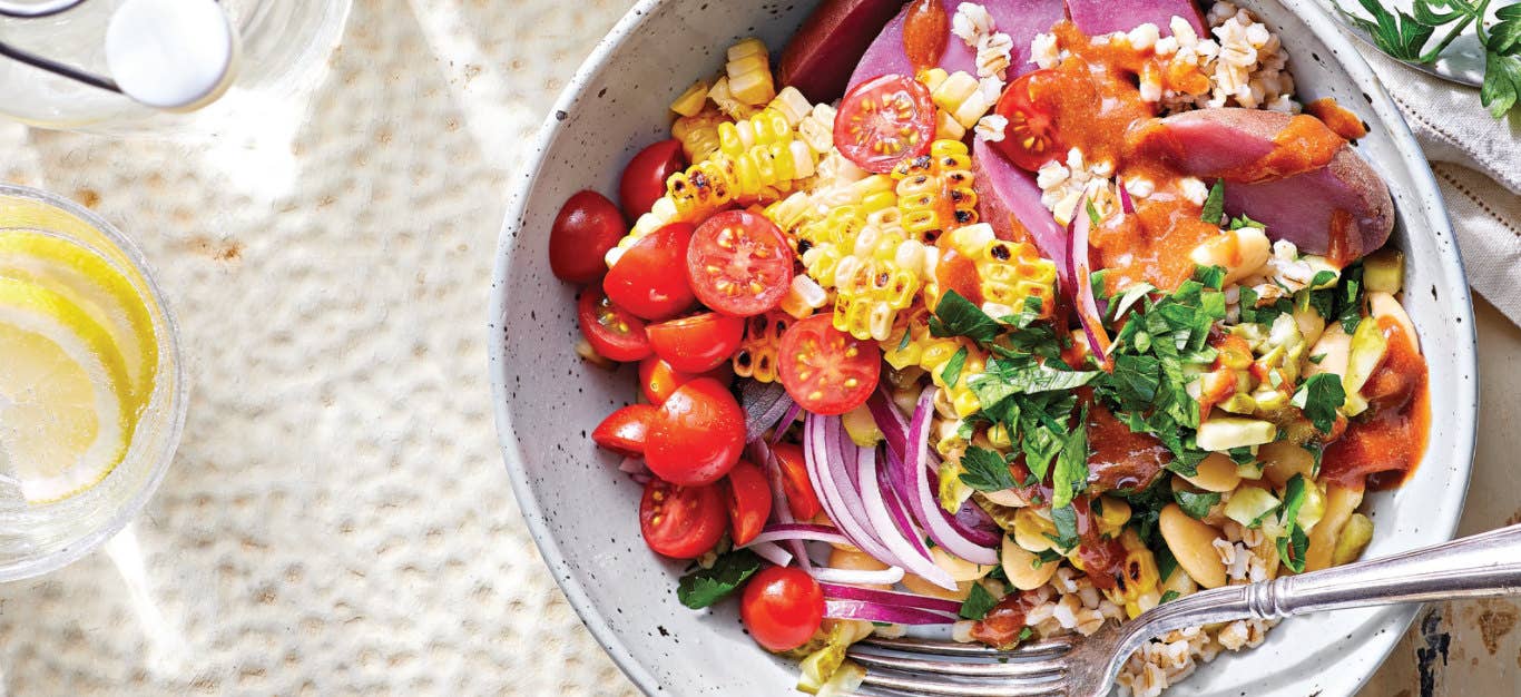 Rainbow Veggie Barley Bowl in a white pottery bowl on a white background next to a small glass of lemon slices