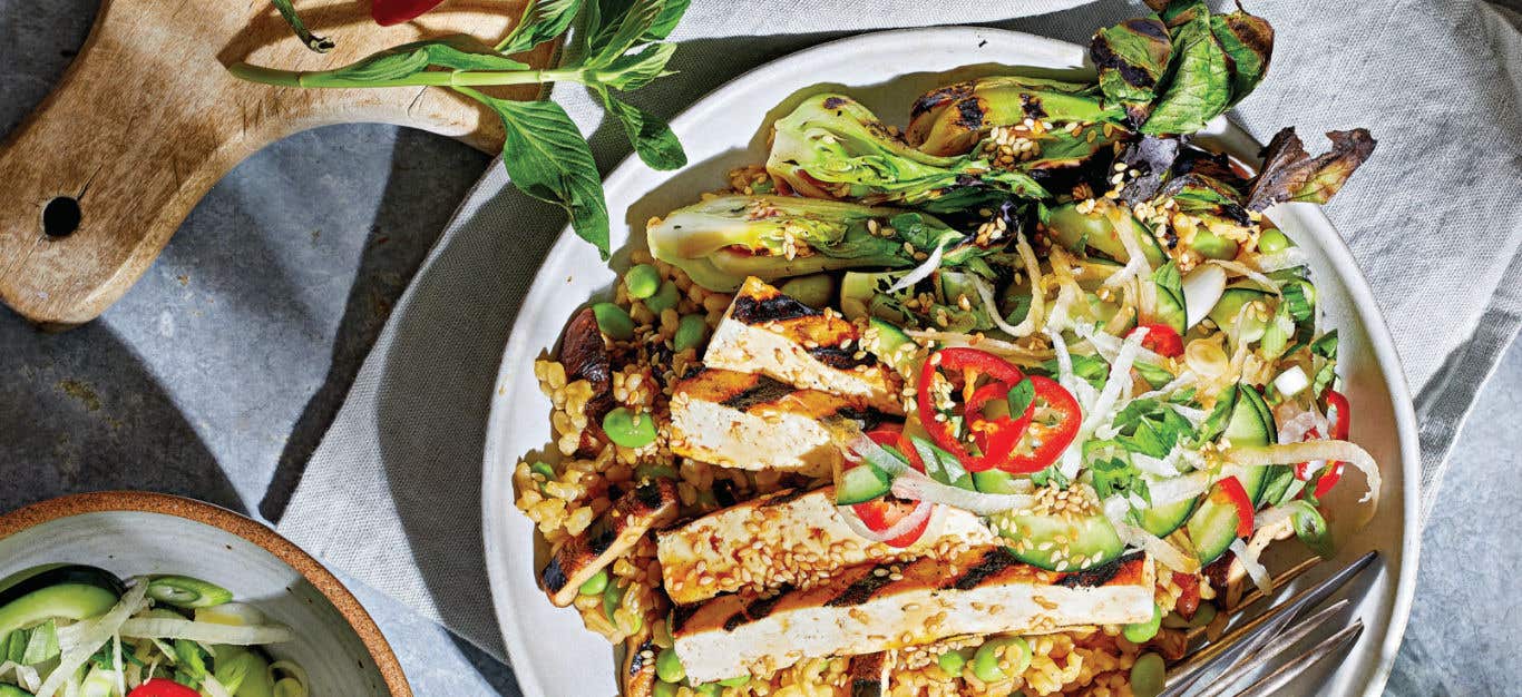 Grilled Bok Choy, Tofu, and Mushrooms with Brown Rice and Ponzu Sauce on a white plate with a sprig of Thai basil on a wooden chopping board