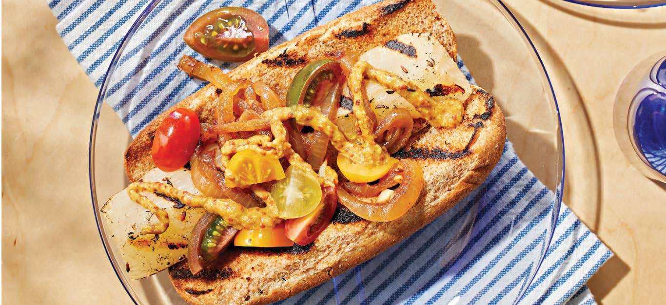 Grilled Veggie Brat on grilled whole grain hot dog bun topped with grape tomatoes, onion, and coarse ground mustard, on a blue and white striped tablecloth.