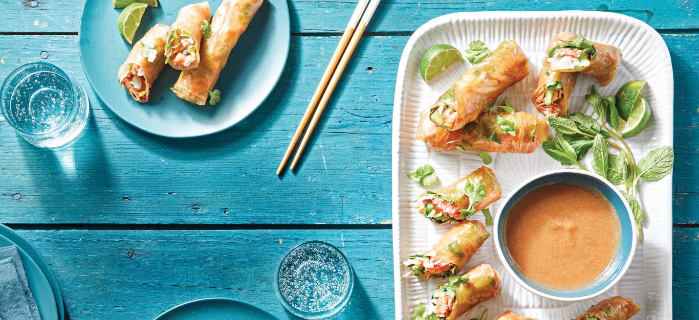 Jackfruit Spring Rolls on a white rectangular serving plate with a small bowl of Sweet-and-Sour Dipping Sauce on a blue wooden table with a smaller plate of spring rolls served up and ready to eat