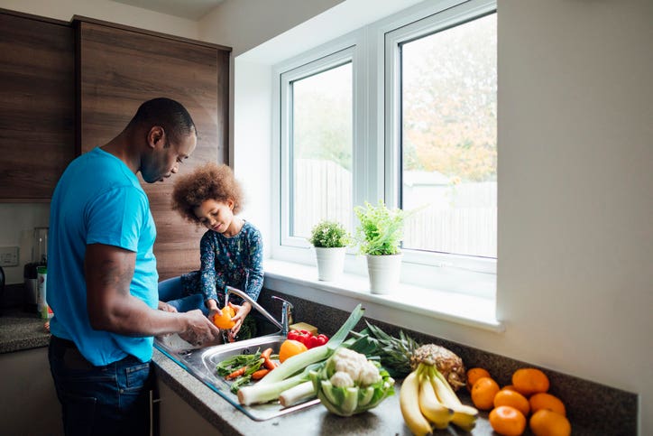 Wide shot of a father and his little girl washing fresh vegetables in the sink at home.