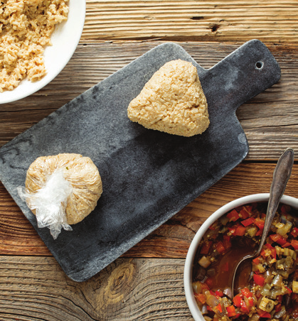 Photo illustrating the following step for making brown rice onigiri: Gently shape the rice into a triangle. Remove the plastic, and sprinkle sesame seeds on top. Repeat Steps 1–4 with remaining rice and filling to form 8 onigiri. 