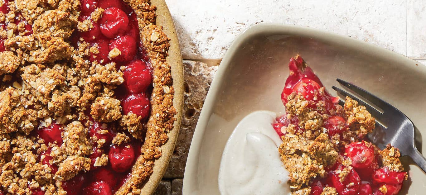 Cherry Crumble Pie in a round brown pie dish next to a plate with a serving of the pie and a dollop of Aquafaba Whipped Cream