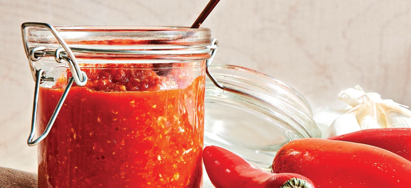 A mason jar of Chile-Garlic Sauce with a teaspoon resting inside, next to red chile peppers and raw garlic