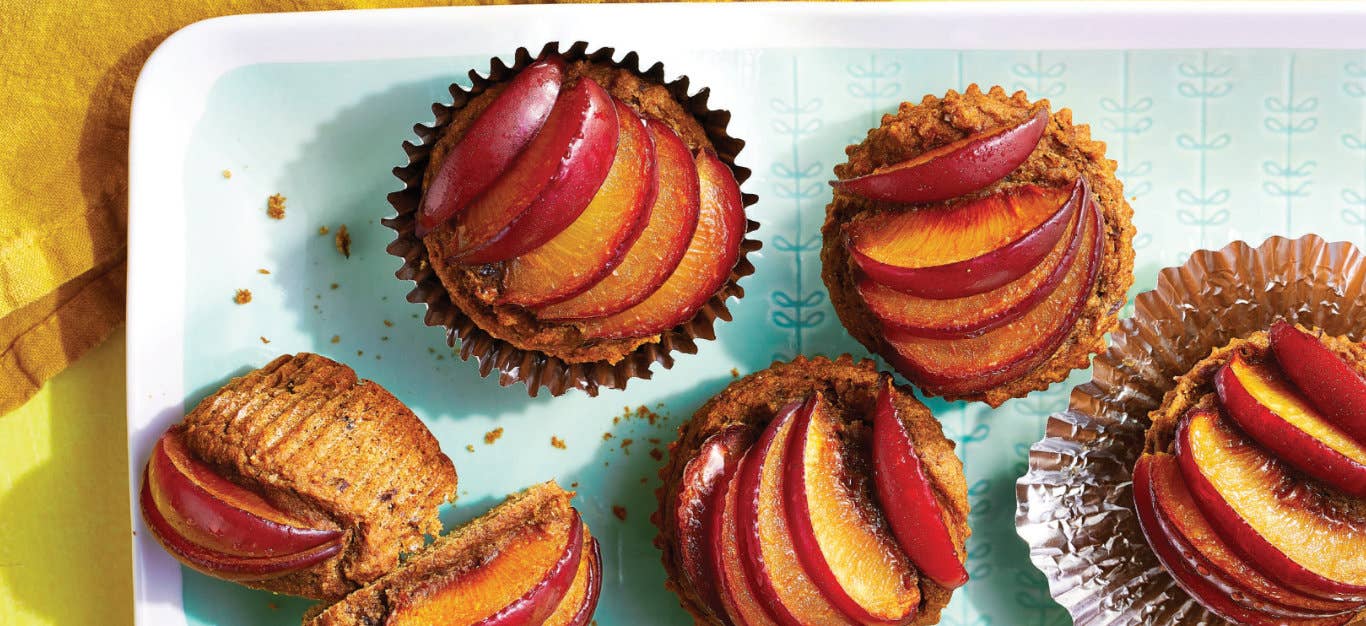 Fresh Plum Muffins in foil muffin papers on a pale blue and white plate with plum slices fanned across the top of each muffin