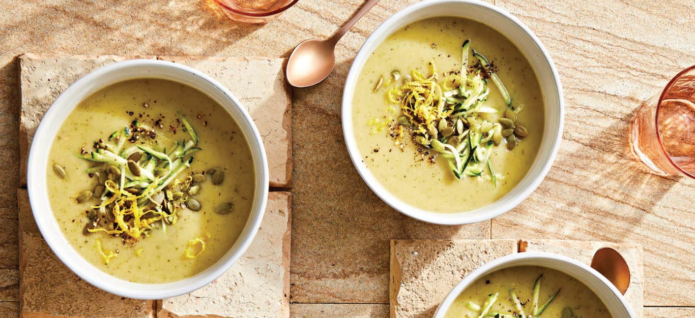 Two white bowls of Creamy Zucchini and Potato Soup with Lemon garnished with lemon zest, pepitas, and grated zucchini