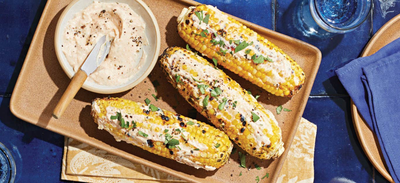 Three cobs of Vegan Elote (Mexican Street Corn) on a platter with a small bowl of lime-infused crema