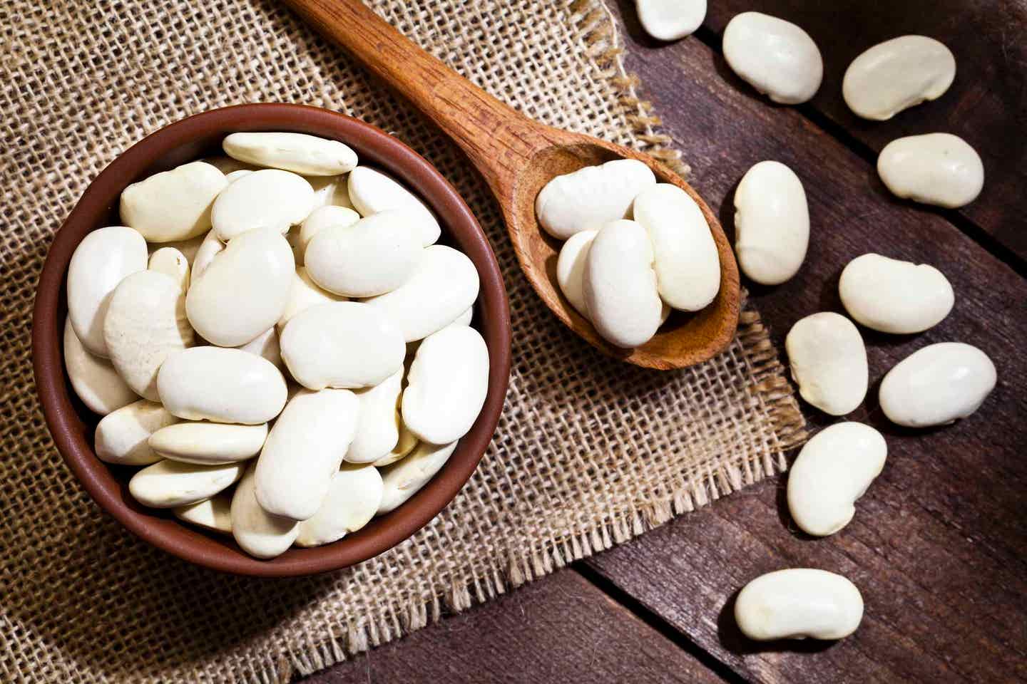 Top view of a brown bowl filled with lima beans shot on rustic wood table. A wooden spoon is beside the bowl with some beans on it.