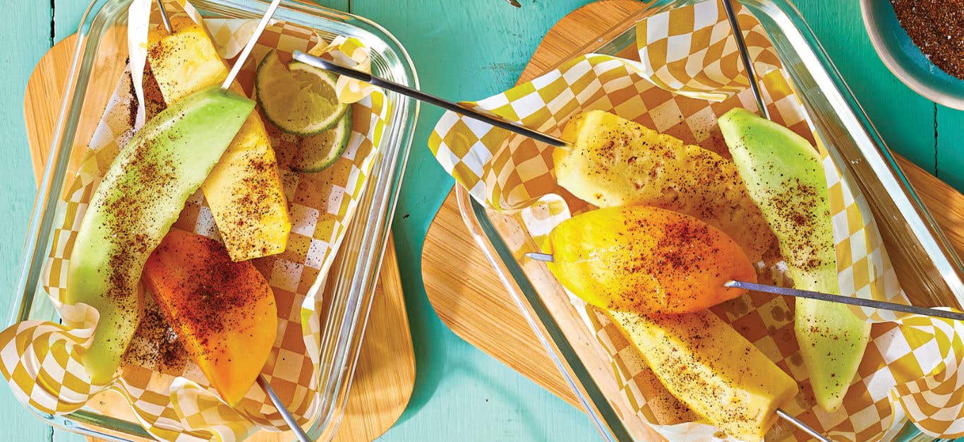 Two servings of Chili-Lime Fruit on a Stick on checked paper napkins in glass Tupperware containers