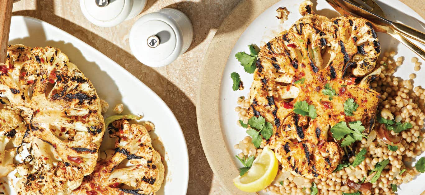 Harissa Cauliflower Steaks with Date Couscous on a white plate next to a salt and pepper shaker and a plate of cauliflower steaks