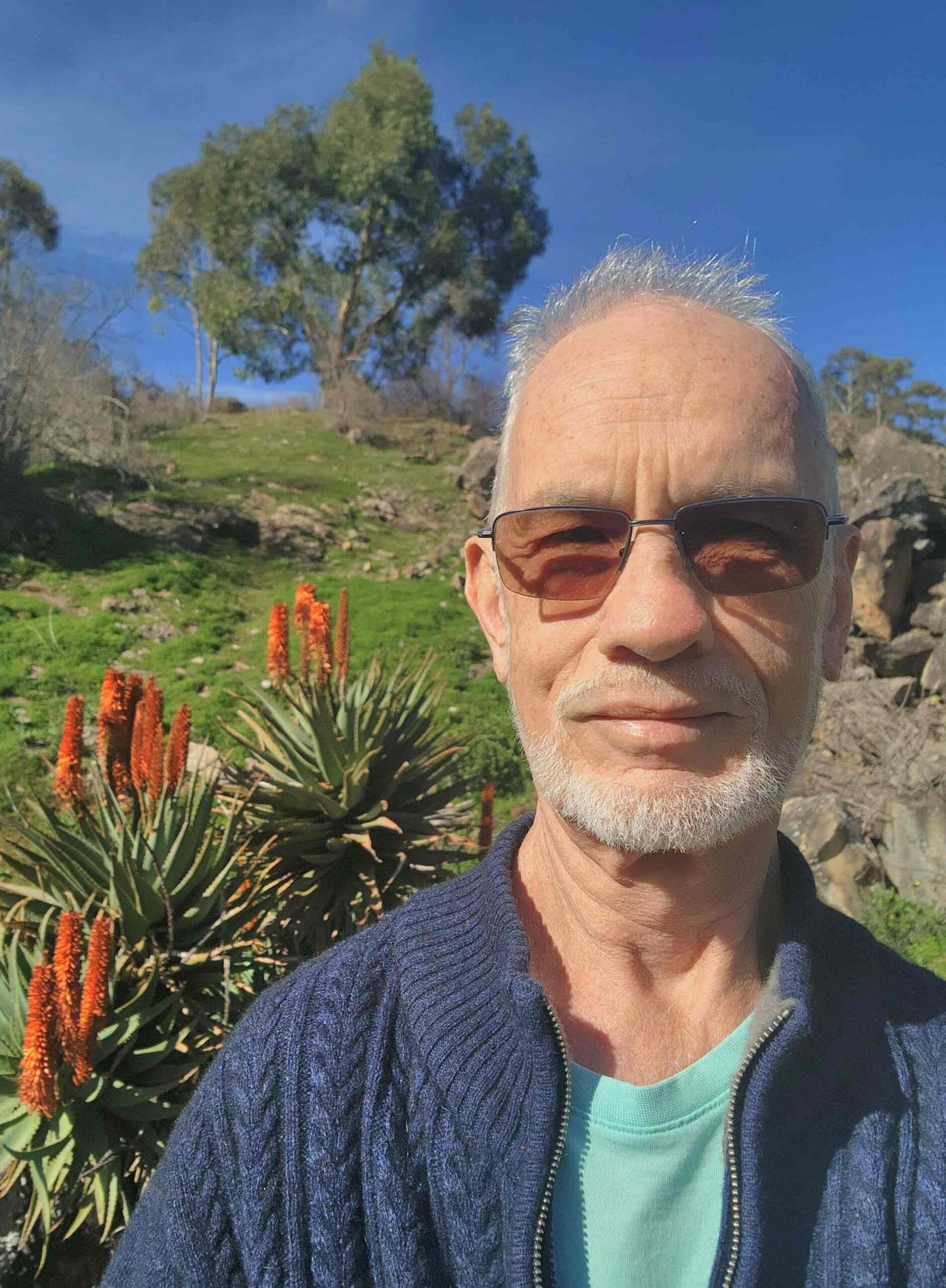 Rainer Lucks, a man in his 60s who adopted a whole food plant based diet for heart disease and got off statins, poses outside, with blue sky and flowers behind him