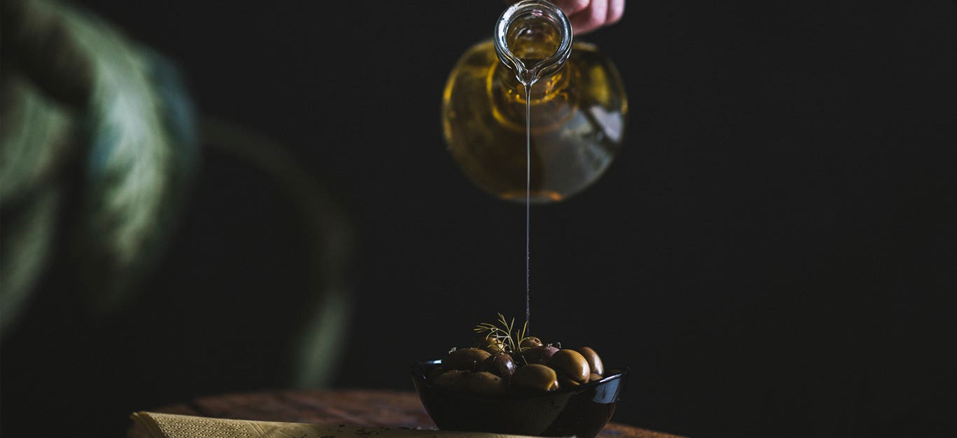 olive oil being poured from a carafe into a small bowl of olives, with a dark background