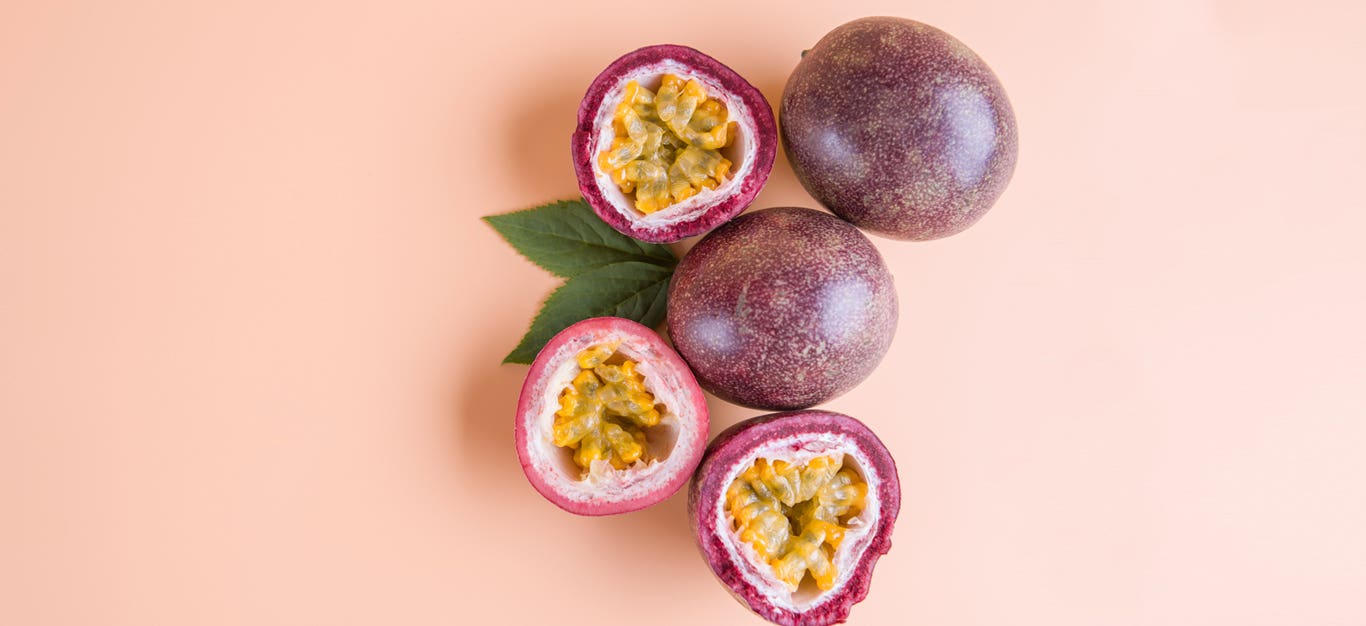 Two whole passion fruit beside three cut-open halves of passion fruit showing the colorful pulp inside, on top of a plain light pink background