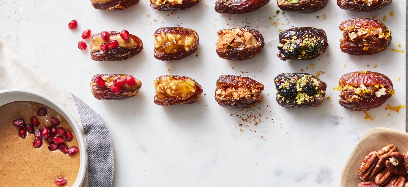 An array of stuffed dates, stuffed with five different fillings, with a small bowl of nut butter and pomegranate arils shown off to the side