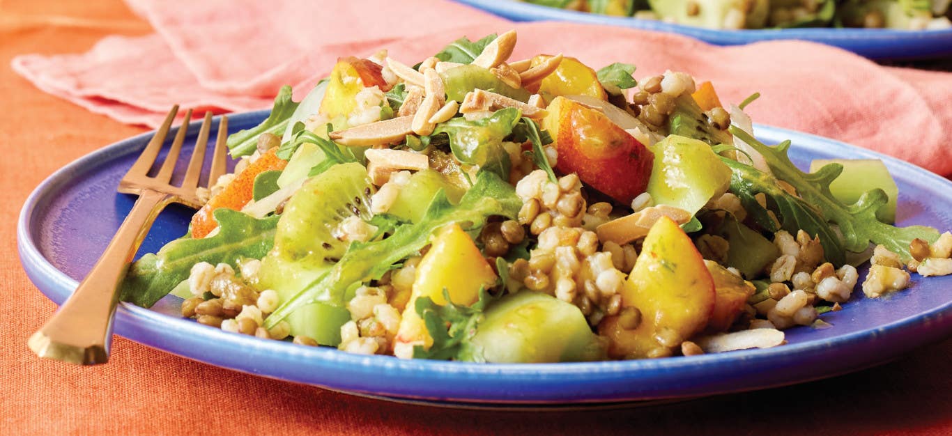 A serving of Honeydew, Peach, and Barley Salad on a blue dinner plate on an orange table top with a pink cloth napkin