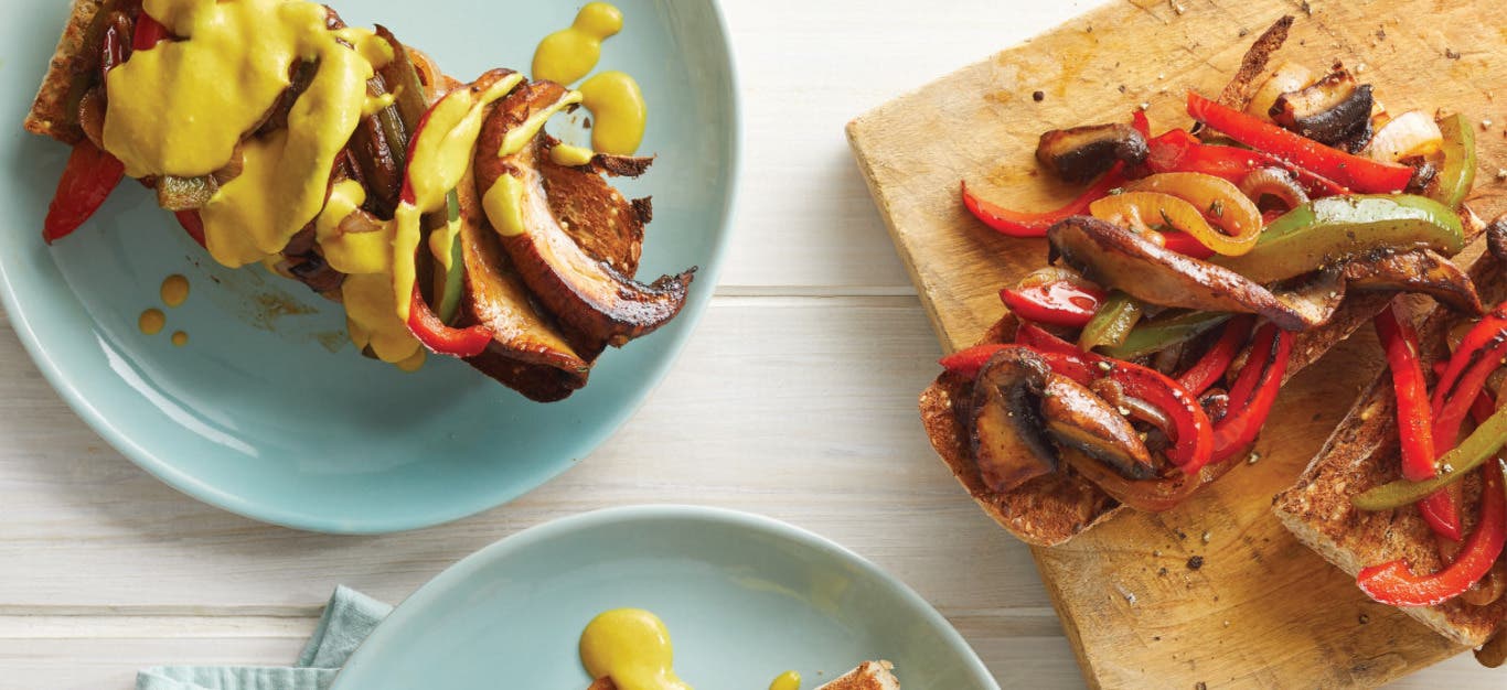 Mushroom Cheesesteak Sandwich on a ceramic plate next to a chopping board with two more sandwiches on it on a white wooden table