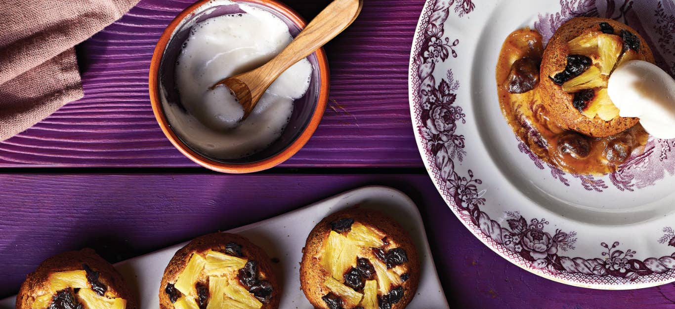 A Upside-Down Pineapple Cupcake on a white and purple tea plate on a purple wooden table next to a small bowl of aquafaba and the corner of a muffin tray showing three cupcakes