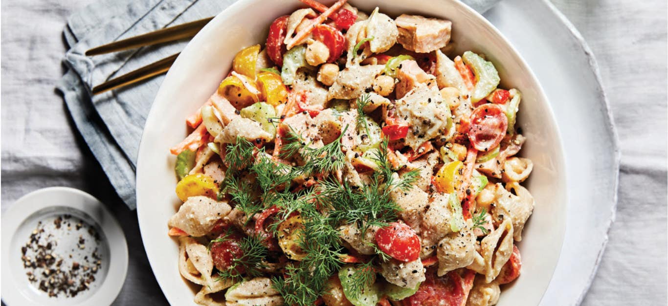 Shellroni Salad with Jackfruit, Cherry Tomatoes, and Creamy Cashew Dressing in a white bowl sitting on a white plate on a white tablecloth, with a garnish of fresh dill on top