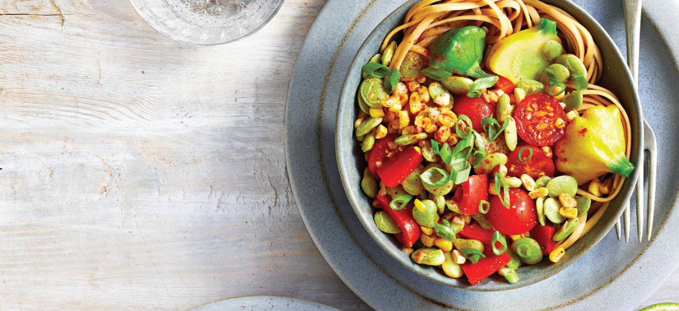 Summer Squash Succotash with Noodles in a blue-grey ceramic bowl sitting on a plate of the same color with a fork on the side