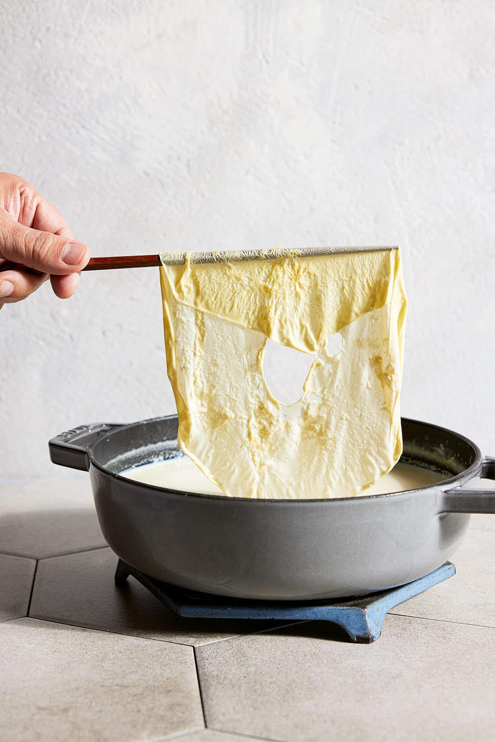 a hand using a chopstick to remove the skin from a pot of homemade soy milk