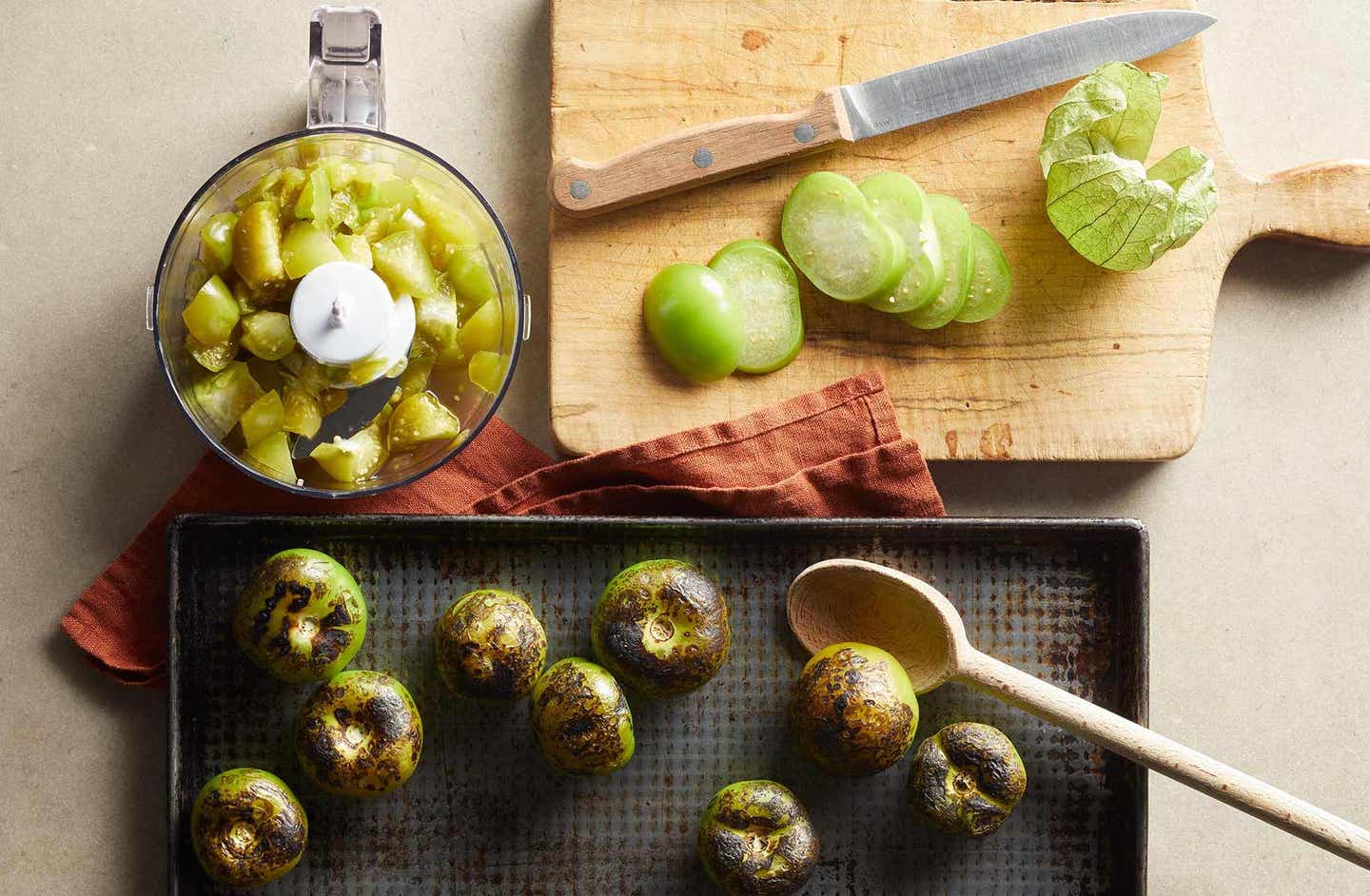 overhead shot of tomatillos chopped in a food processor, beside a cutting board with a sliced tomatillo and a knife, with broiled roasted tomatillos on a pan below