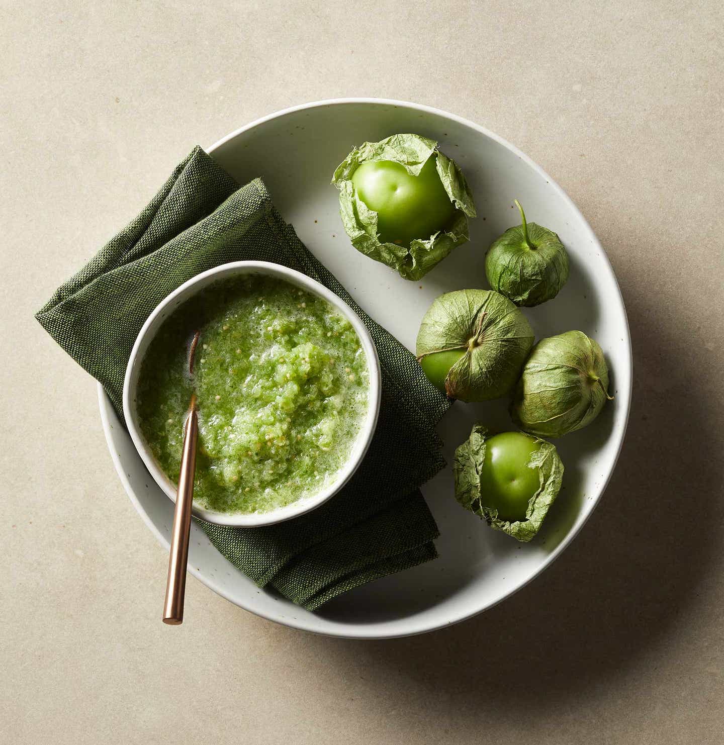 A bowl of bright green salsa verde, on a plate with whole tomatillos next to it