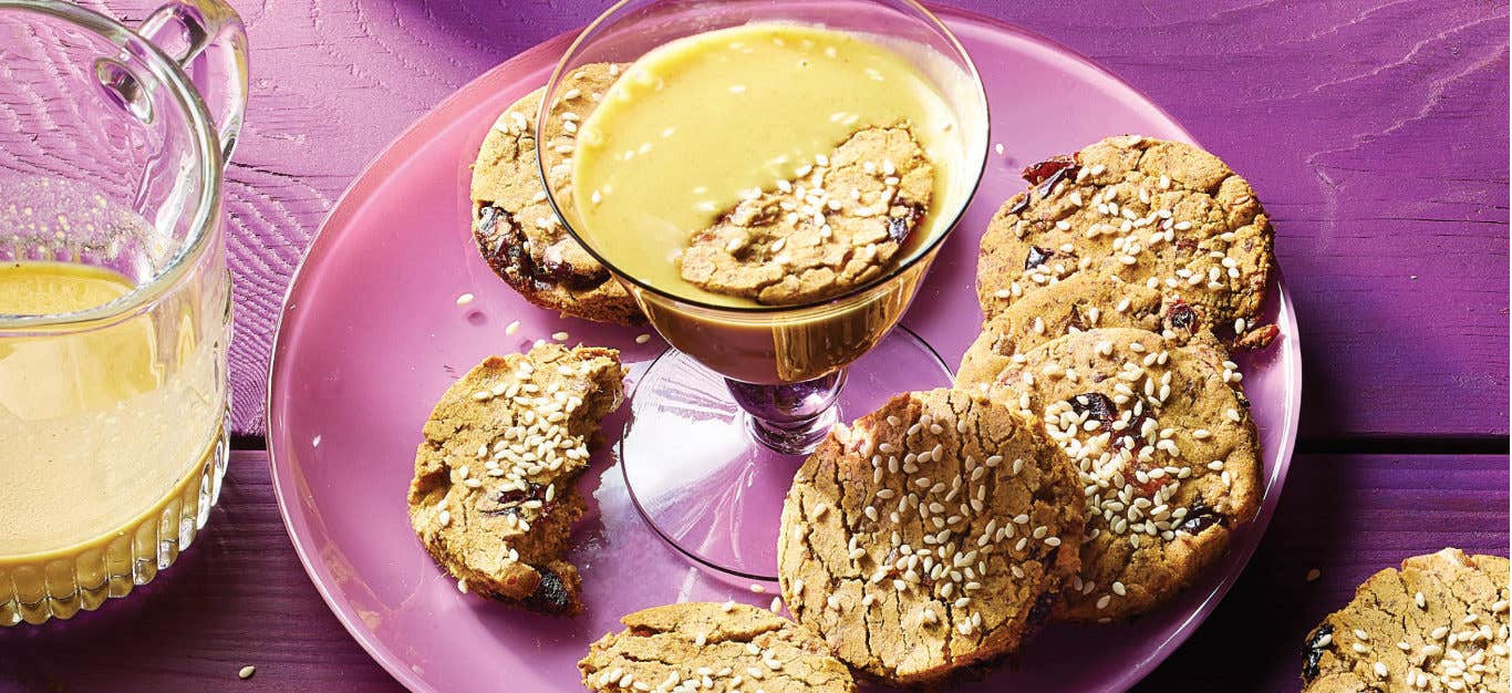 Sesame Date Cookies on a pink plate with a long stemmed glass filled with Golden Turmeric Milk in the middle of the plate