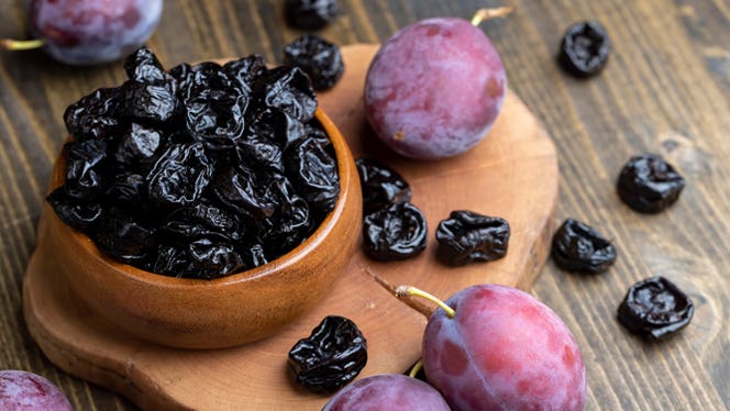 a bowl of prunes shown next to a few fresh plums on a wooden surface