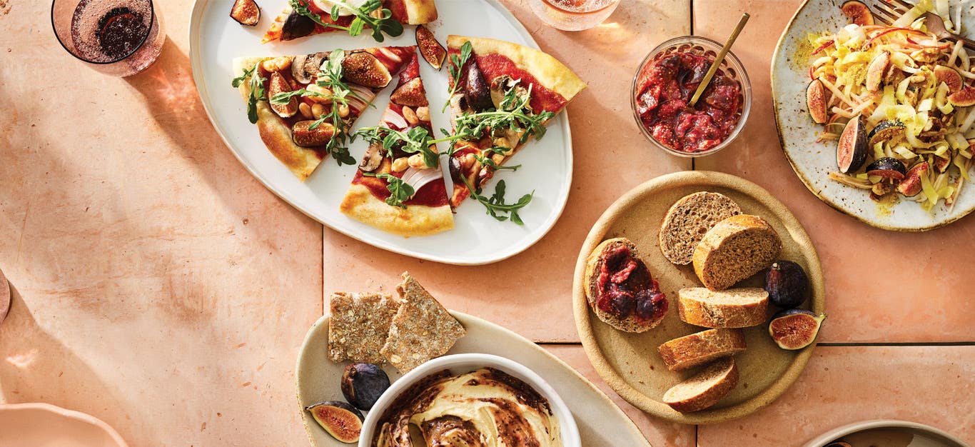 A table spread with a variety of fresh fig dishes: a pizza topped with fresh figs, hummus in a ramekin with a fig paste swirled in, fig jam spread on whole grain bread, a fig and apple salad, and maple-roasted figs