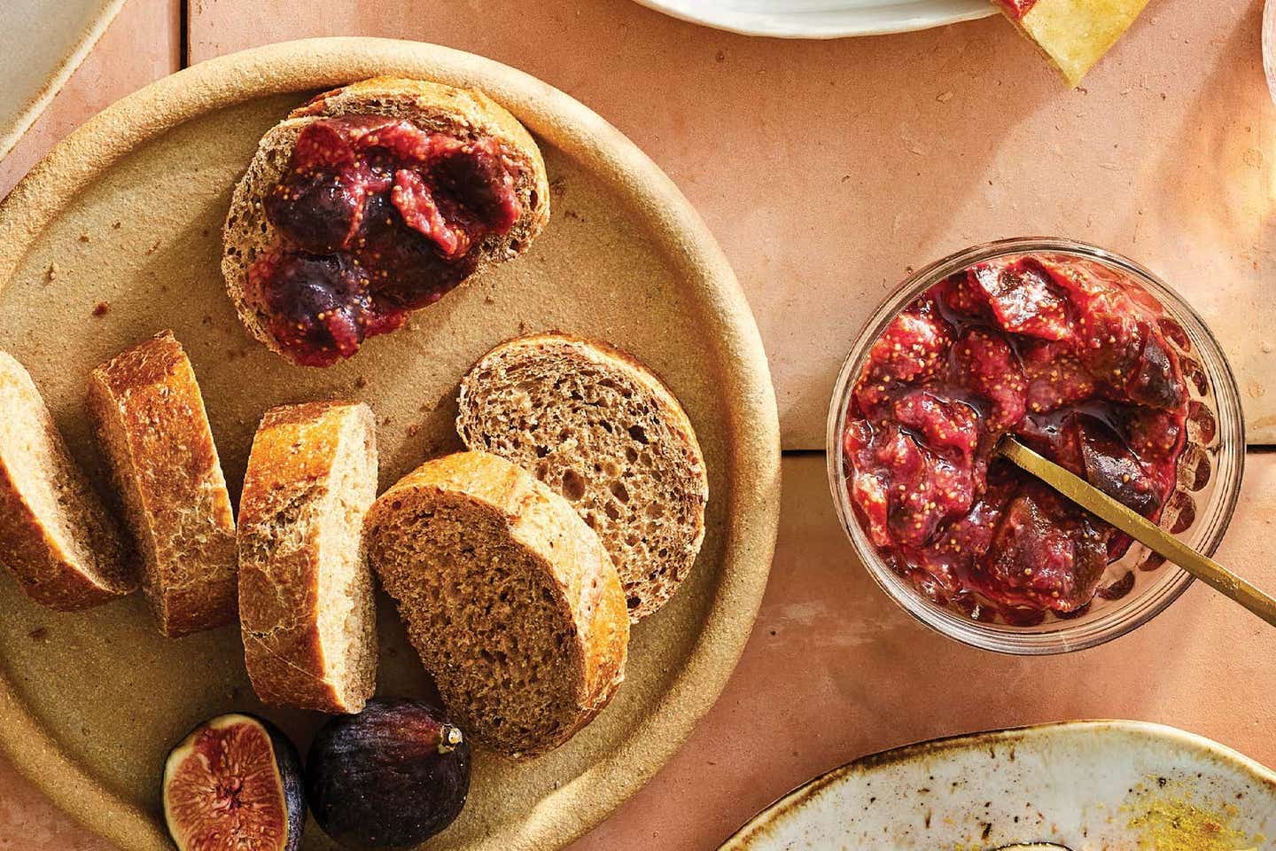 Fig jam with ginger, shown in a jar next to a few slices of whole grain bread, with jam spread on one slice of the bread