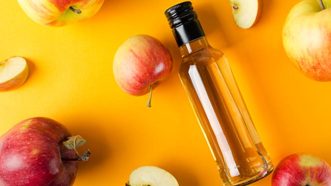 A bottle of apple cider vinegar lays on an orange background beside red apples