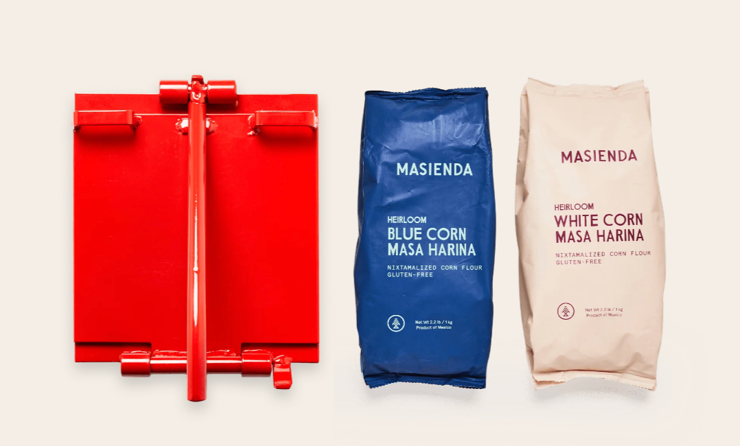 The Masienda-brand tortilla starter kit, with a bright-red tortilla press and two bags of masa harina (blue corn and white corn)