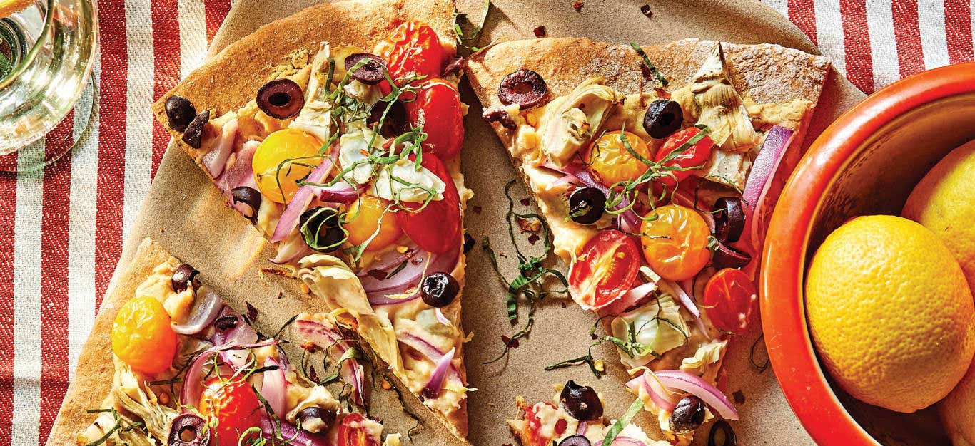 Mediterranean Artichoke Pizza on a light-weight wooden platter next to a small red bowl of lemons, with a red and white striped dish towel behind