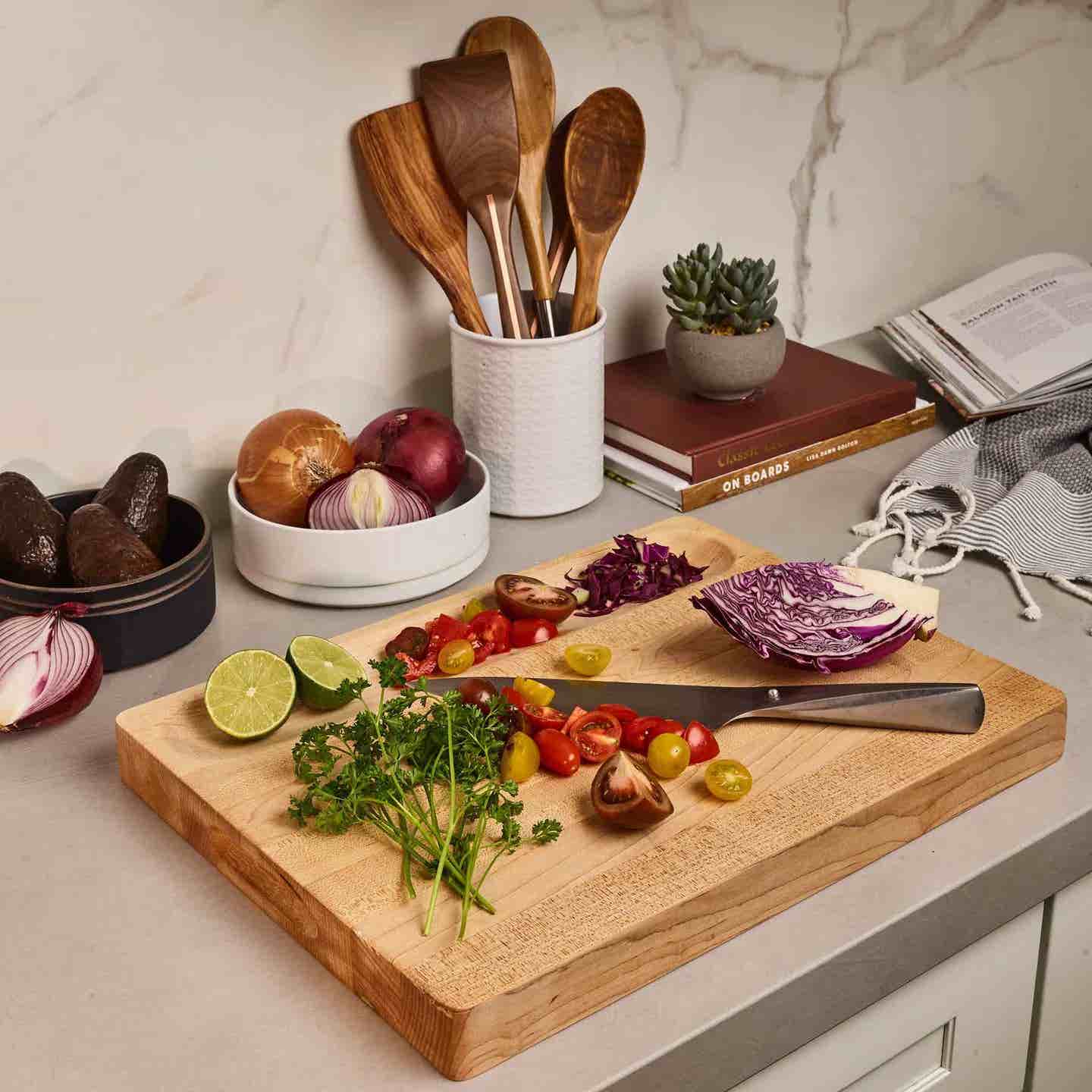 The WInsome Maple Cutting Board by the brand Sonder. A wooden cutting board sits on a counter; on the board are chopped cherry tomatoes, fresh herbs, a lime, with some of the chopped ingredients sitting inside a divot along one side of the cutting board