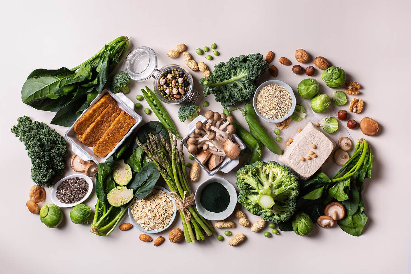 a wide array of protein-rich whole plant foods shown arranged on a white surface, including tofu, broccoli, tempeh, nuts, and seeds