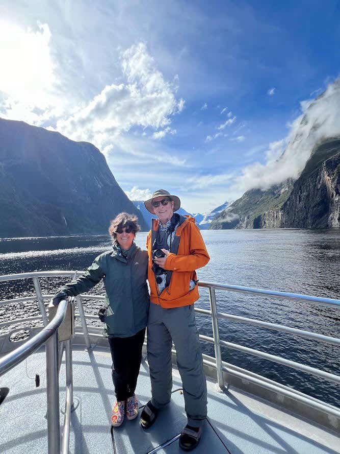 A woman and man in their 60s stand on the front of a large boat with a beautiful lake and blue sky in the background, wearing hiking clothing